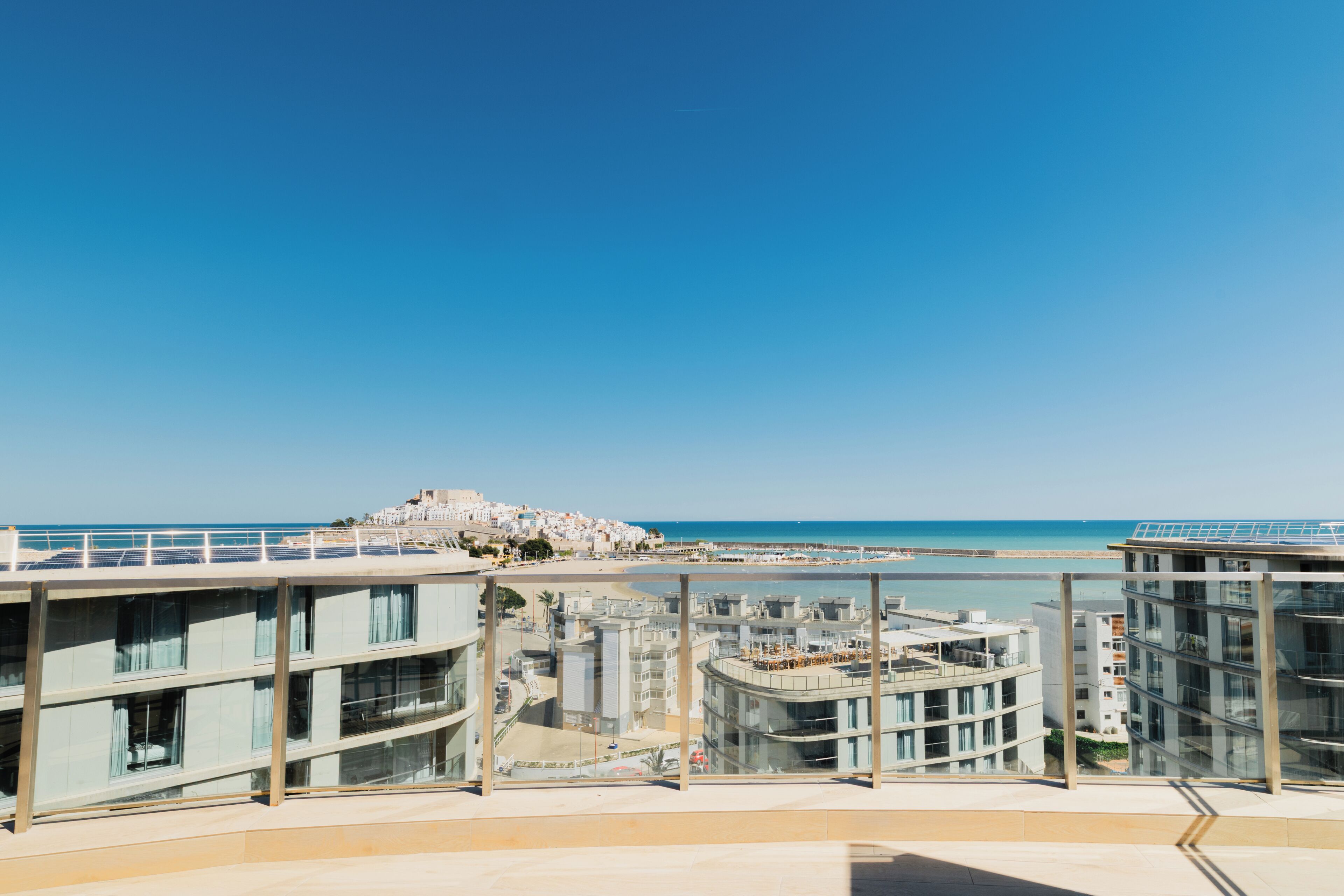 A modern building with a glass facade, a balcony with a view of the beach, and a clear blue sky.