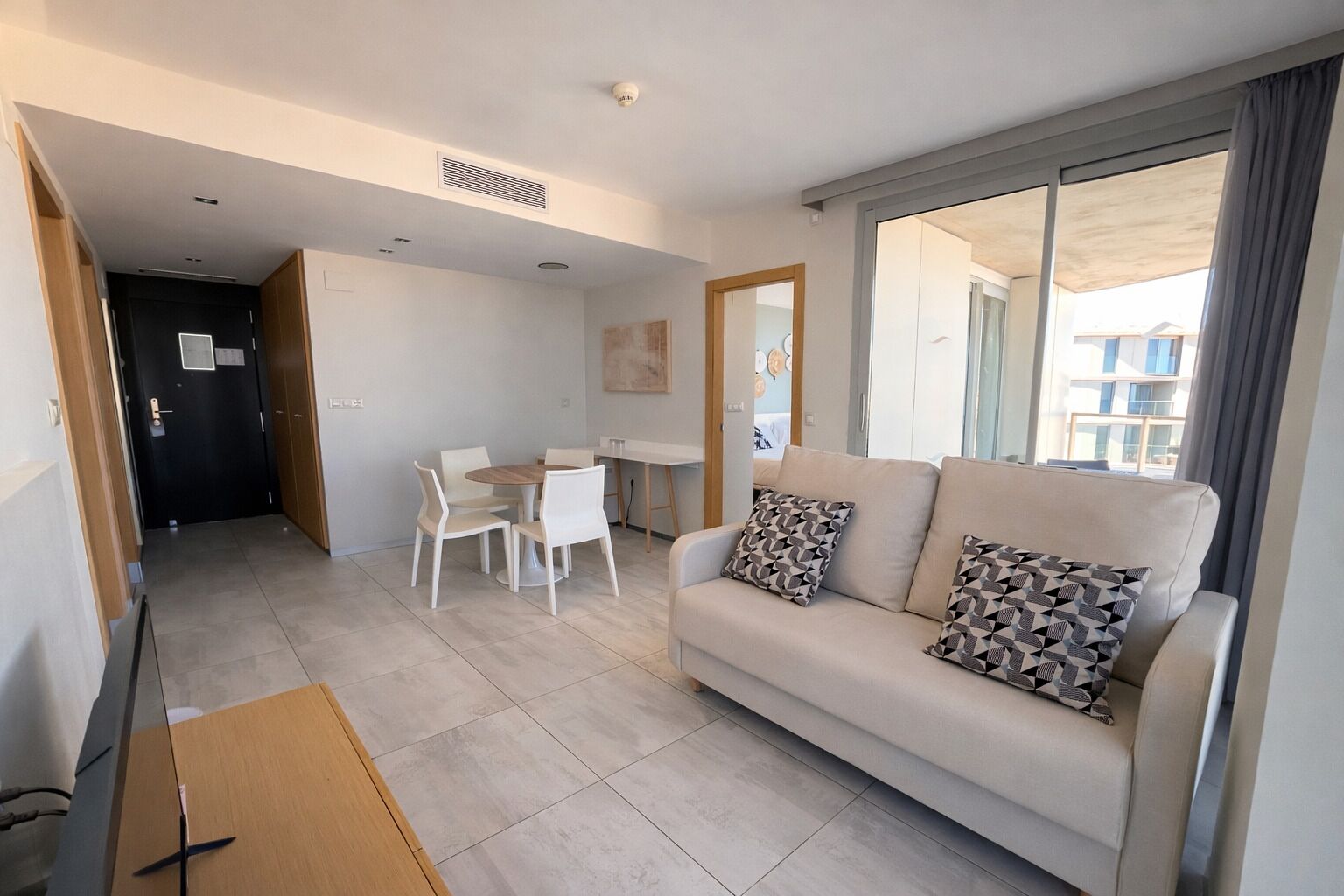 Una sala de estar moderna con un sofá, dos cojines estampados, una mesa de comedor con sillas y vistas a través de un ventanal.