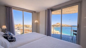 Habitación de hotel con dos camas, un balcón que ofrece vistas al mar y al paisaje urbano, y una lámpara.
