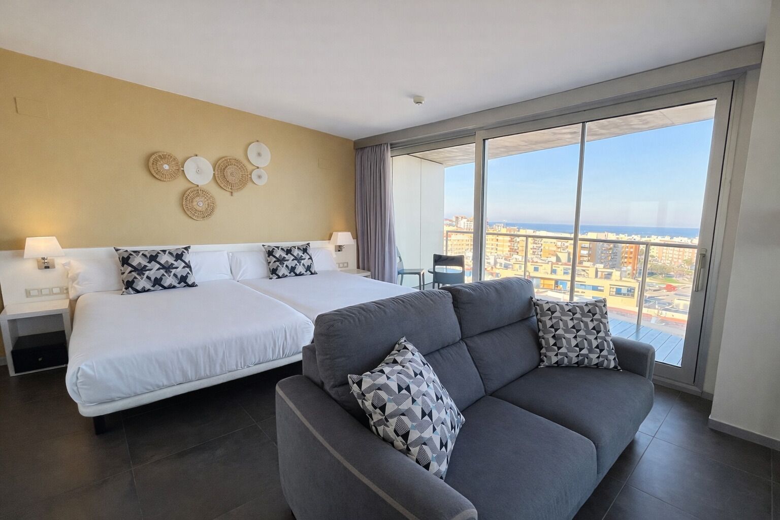 A hotel room with two beds, a grey sofa, and a large window offering a city view.