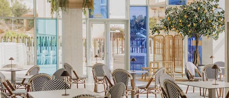 A spacious indoor area with multiple tables and chairs, large windows, and a large tree with yellow fruits.