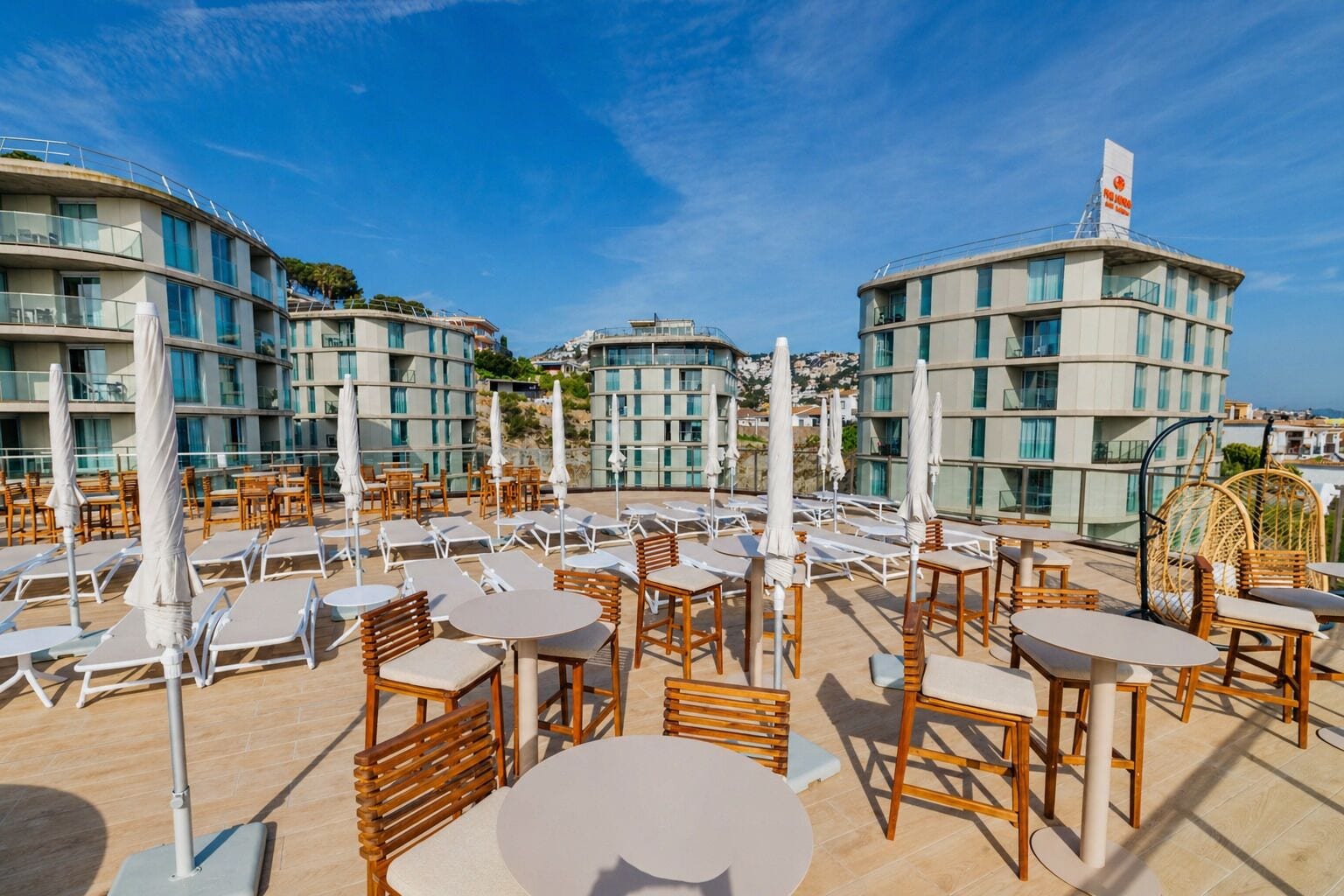 Terraza en la azotea con sillones de sol, mesas y sombrillas, rodeada de edificios modernos con balcones.