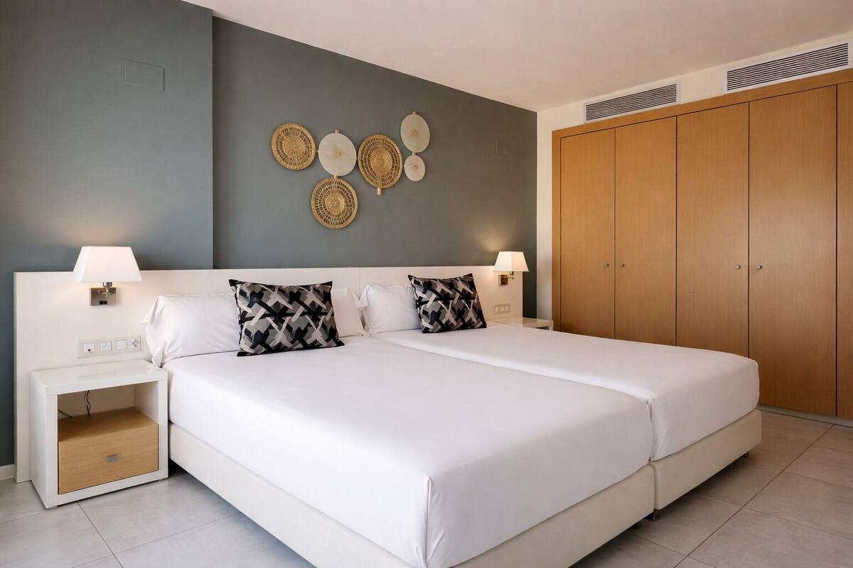 Una cama doble con ropa de cama blanca, dos mesitas de noche con lámparas, un adorno en la pared y un armario de madera.