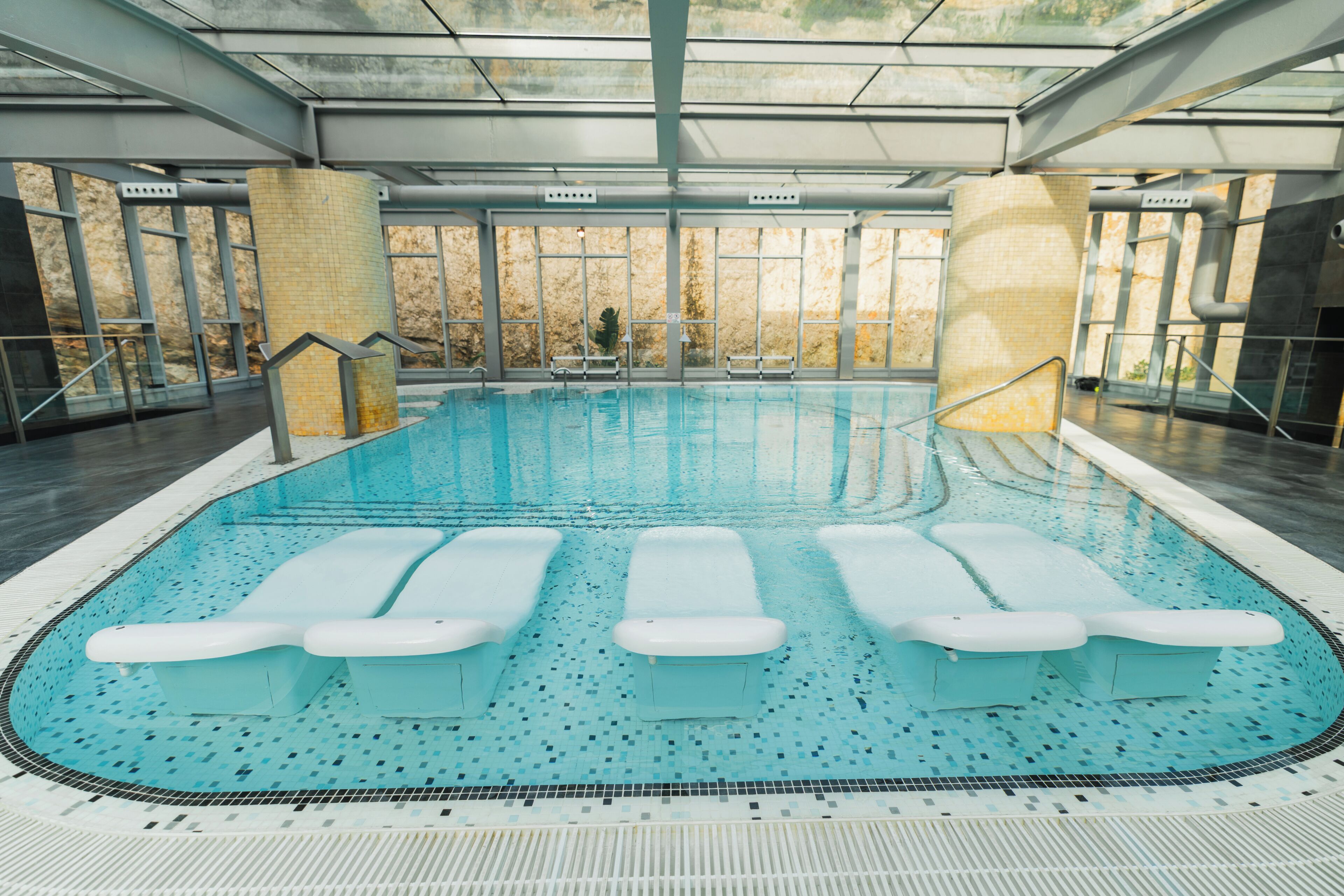 Ein großer, überdachter Pool mit mehreren Liegestühlen, gefliestem Boden und großen Fenstern.