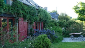 Une maison à l’extérieur rouge, un jardin avec différentes espèces de plantes et une terrasse avec une table et des chaises.