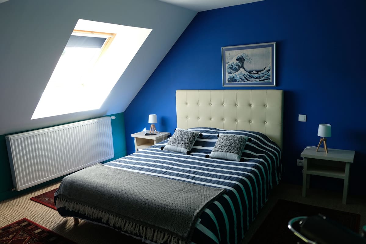 Een slaapkamer met een schuine plafond, een bed met gestreepte beddengoed, een blauwe muur, een lijstwerk met kunst en een dakraam.
