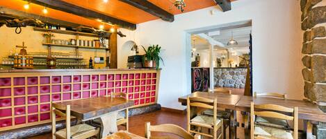 Un interior de restaurante con mesas y sillas de madera, una zona de bar con estanterías y una pared de piedra.