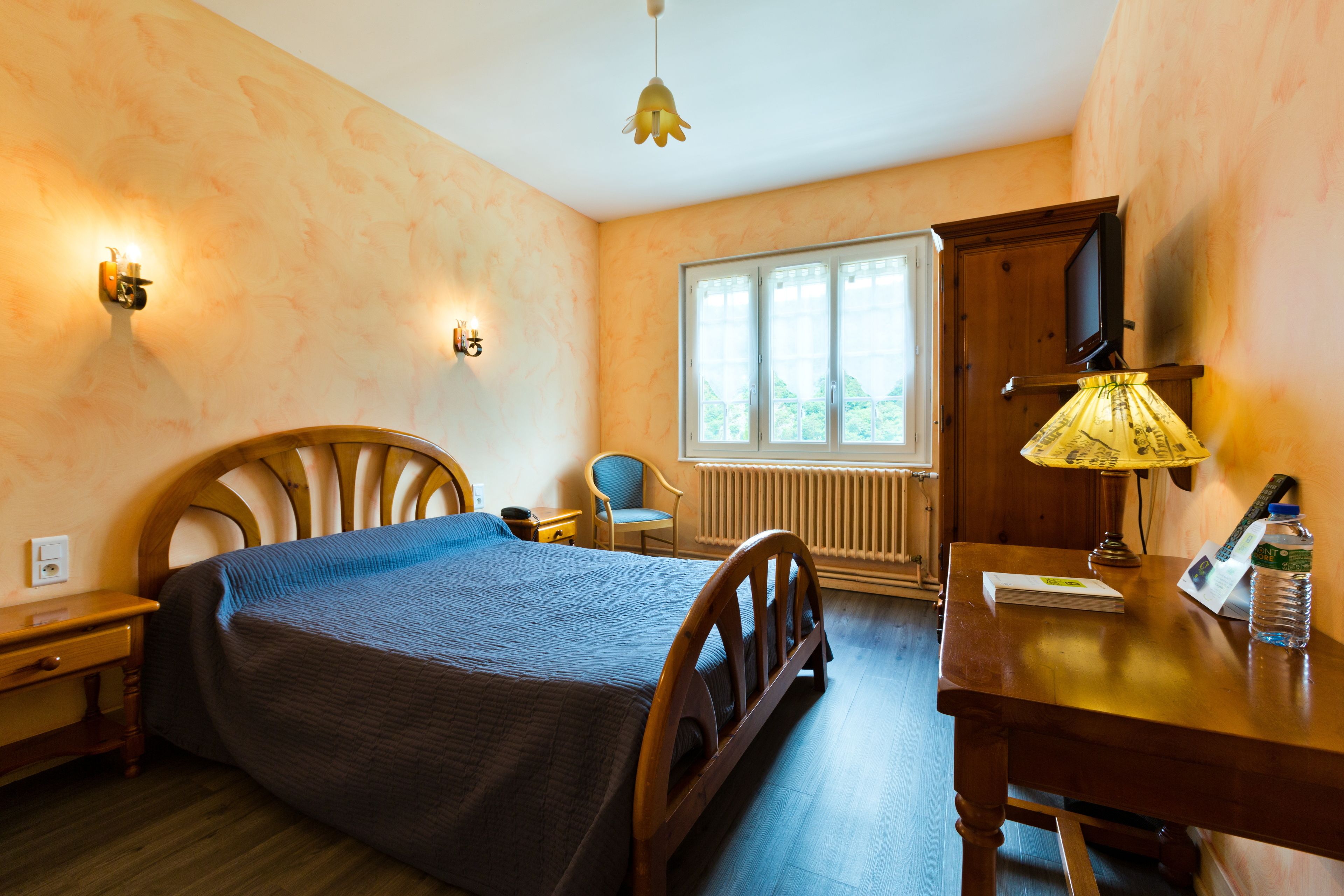 A bedroom with a wooden bed, a desk with a lamp, a chair, and a window with a view of greenery.