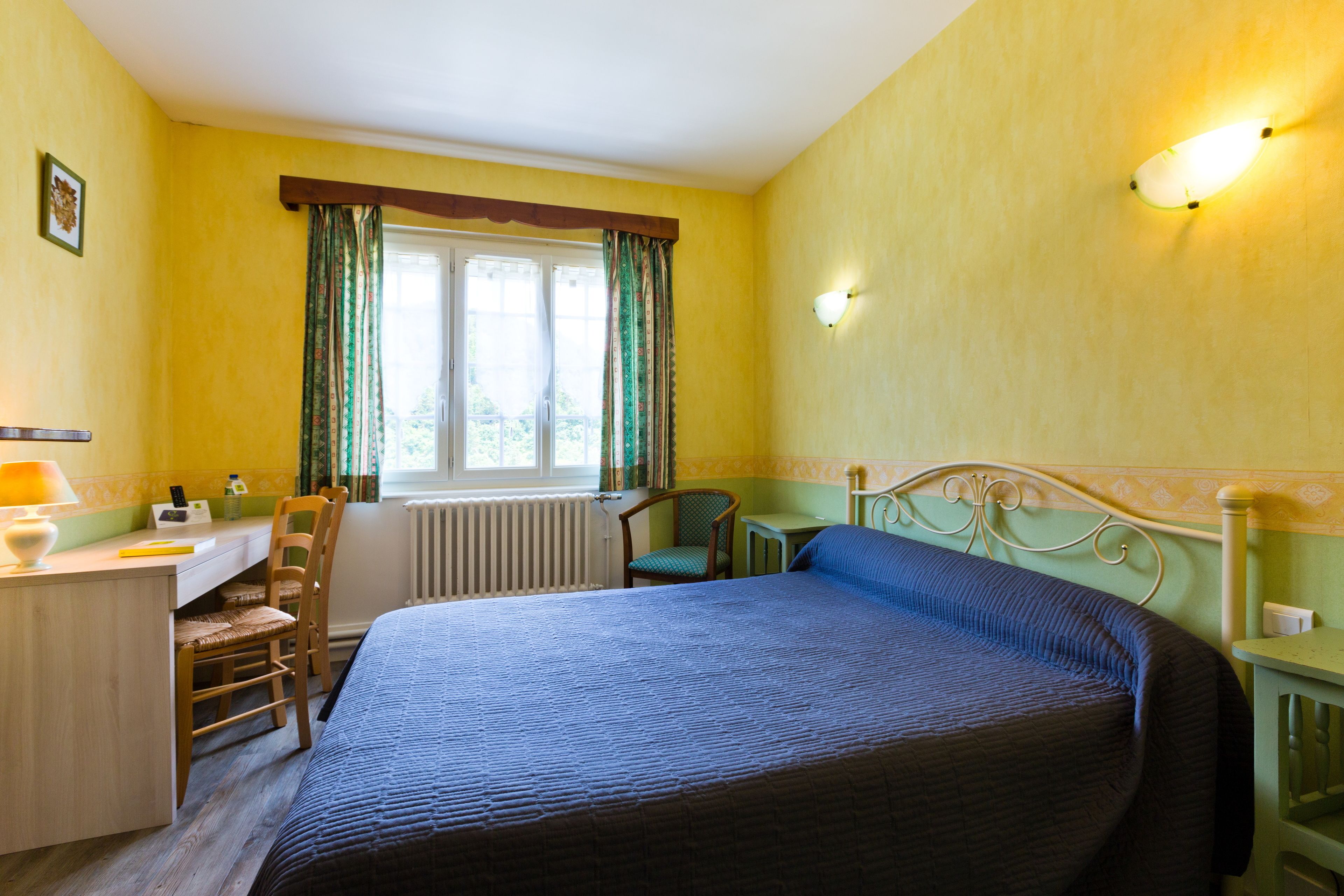 A bedroom with a bed, a desk, a chair, a window with curtains, and a wall-mounted light fixture.