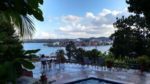 An outdoor pool area with a view of a coastal town, surrounded by greenery and potted plants.