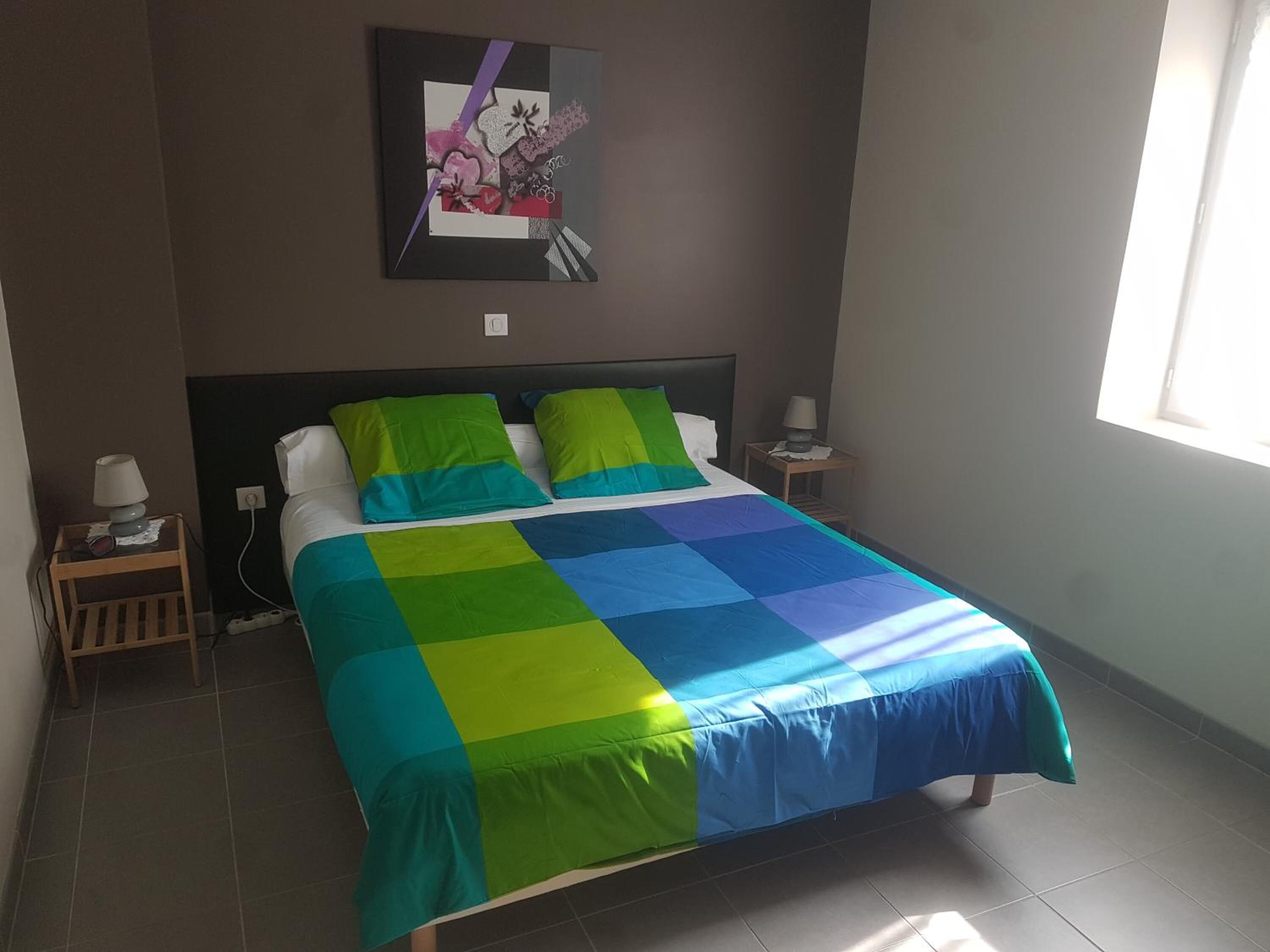 Una cama bien tendida con un colcha colorida, una mesita de noche con una lámpara y una obra de arte en la pared sobre la cama.