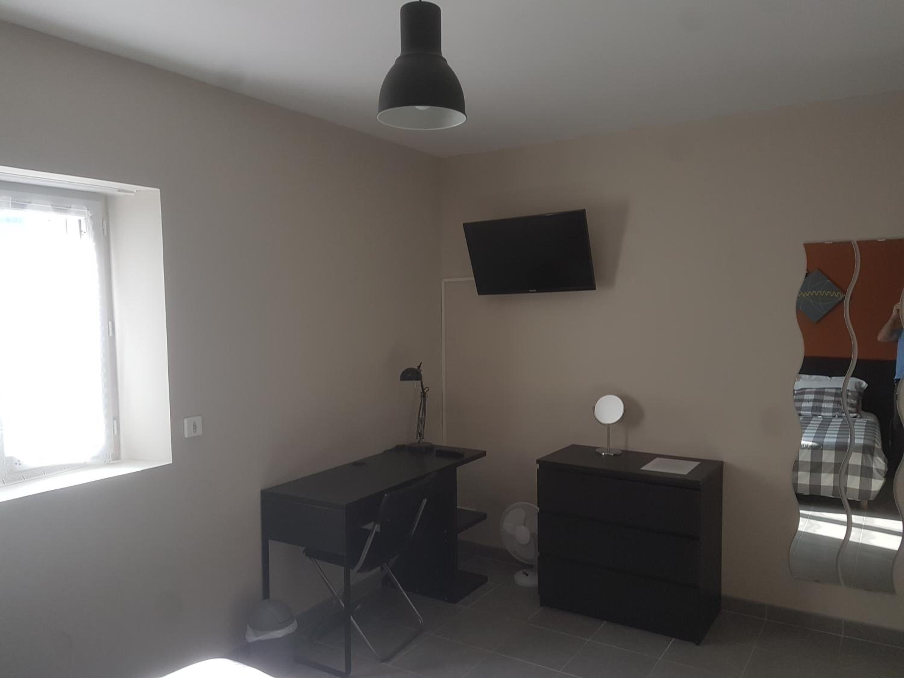 A room with a desk, chair, a small table with a lamp, a TV mounted on the wall, and a bed with a checkered blanket.