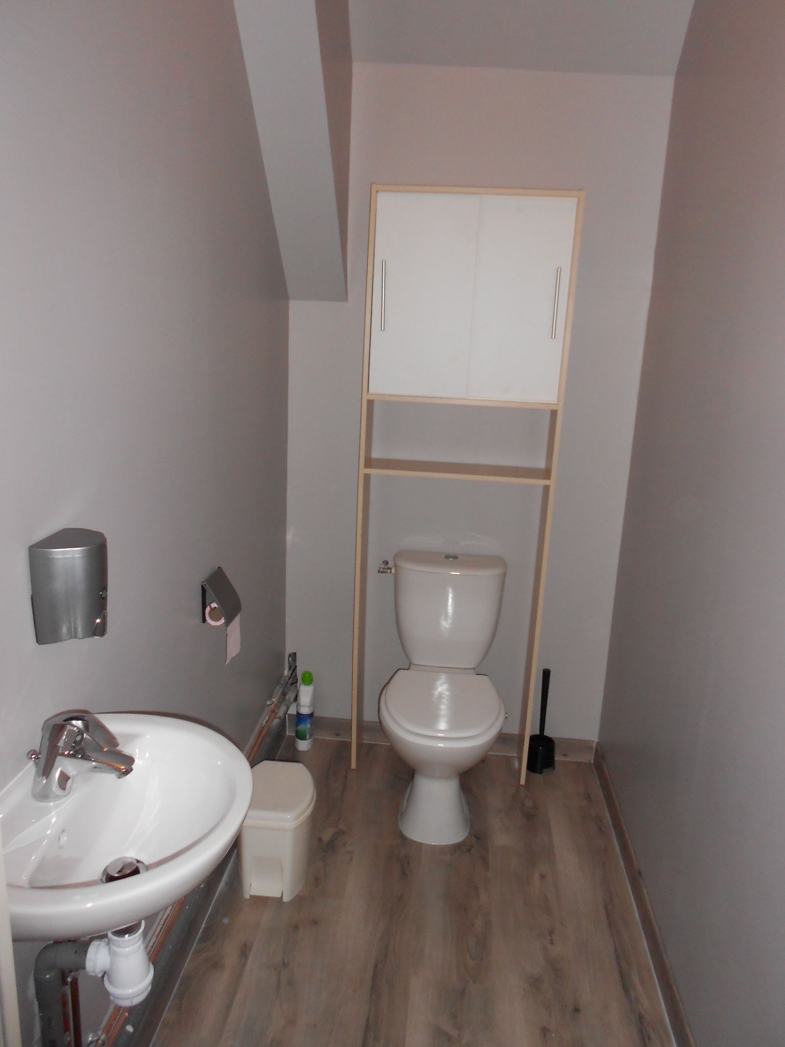 Un baño peque ño con un lavamanos blanco, inodoro y un estante de madera sobre el inodoro.