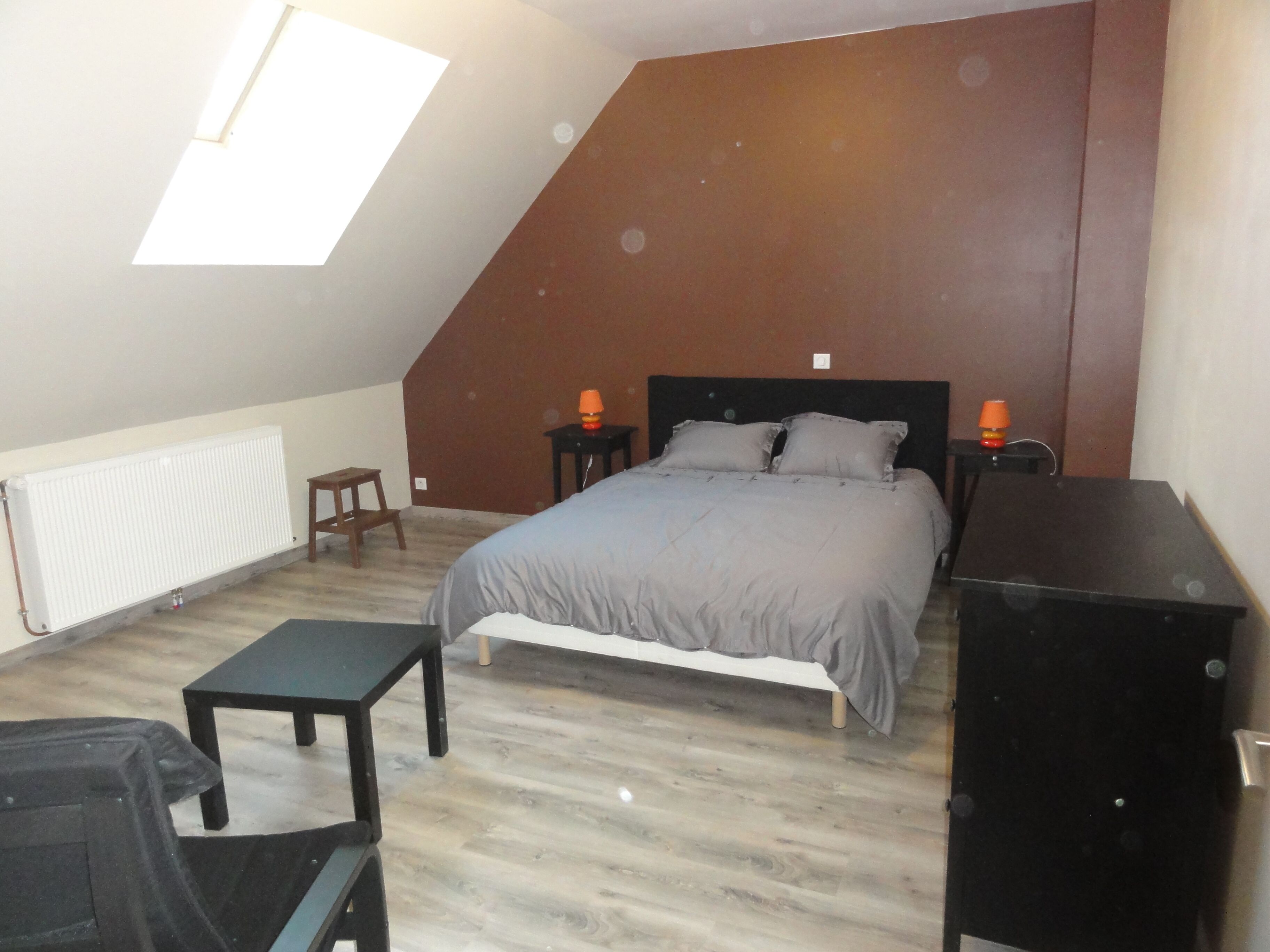 A bedroom with a sloped ceiling, a bed with grey bedding, two bedside tables with lamps, a radiator, a small wooden stool, and a black desk.