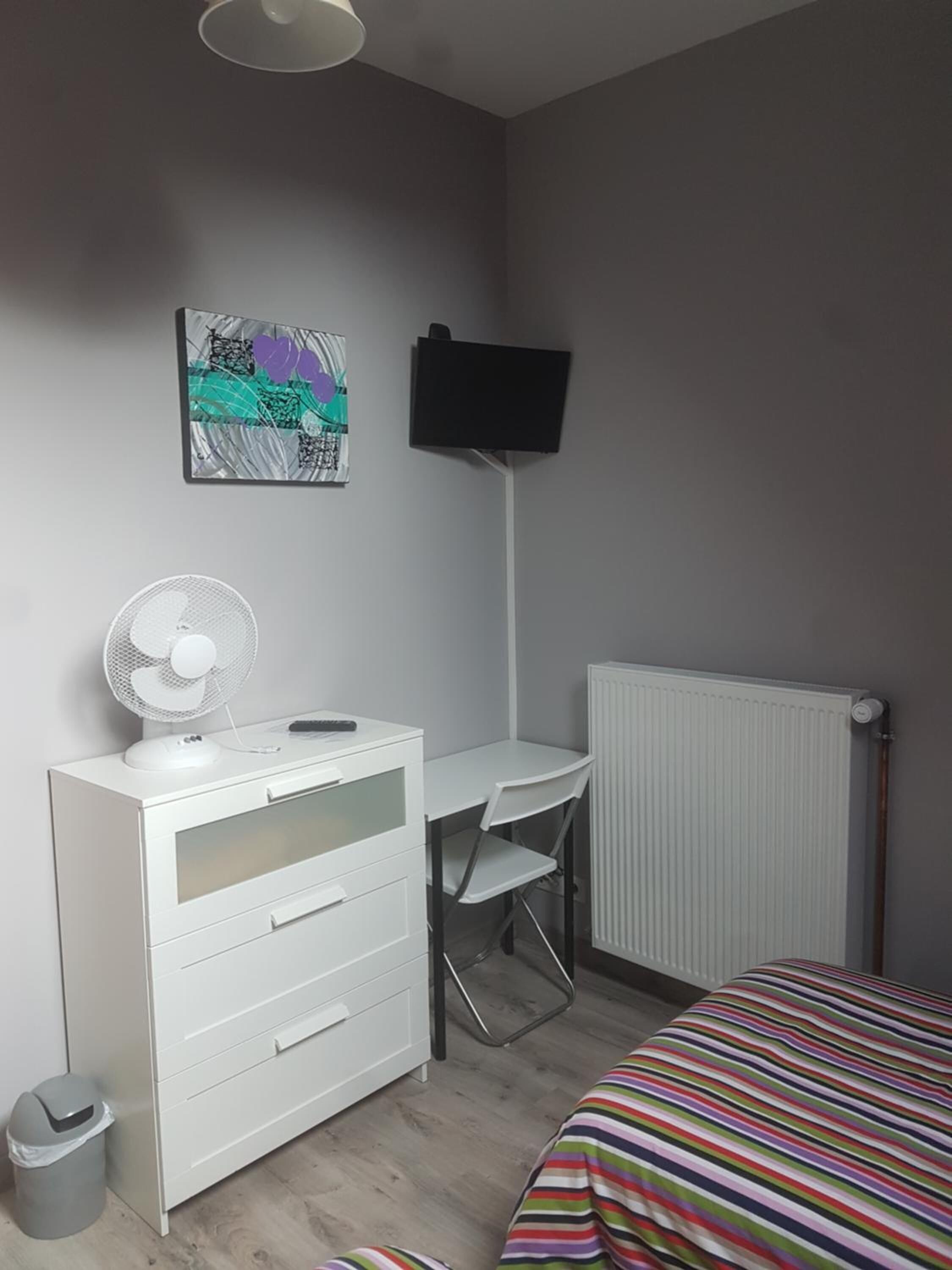 A small, neatly organized bedroom with a bed, a desk, a chair, a radiator, a white dresser, a trash bin, and a wall-mounted TV.