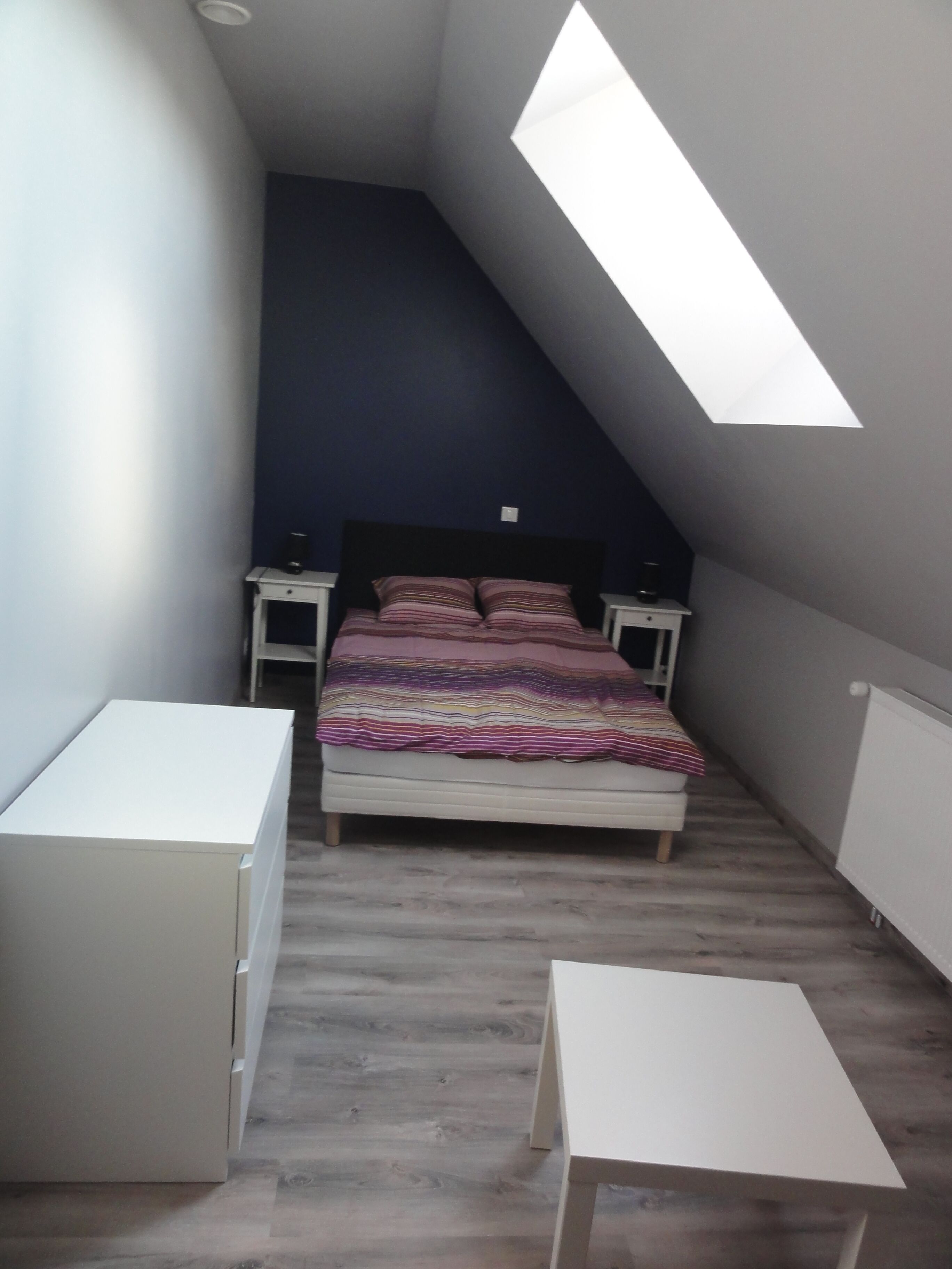Een slaapkamer met een schuine kap, een bed met een gestreept dekbedovertrek, nachtkastjes en een klein witte tafeltje.