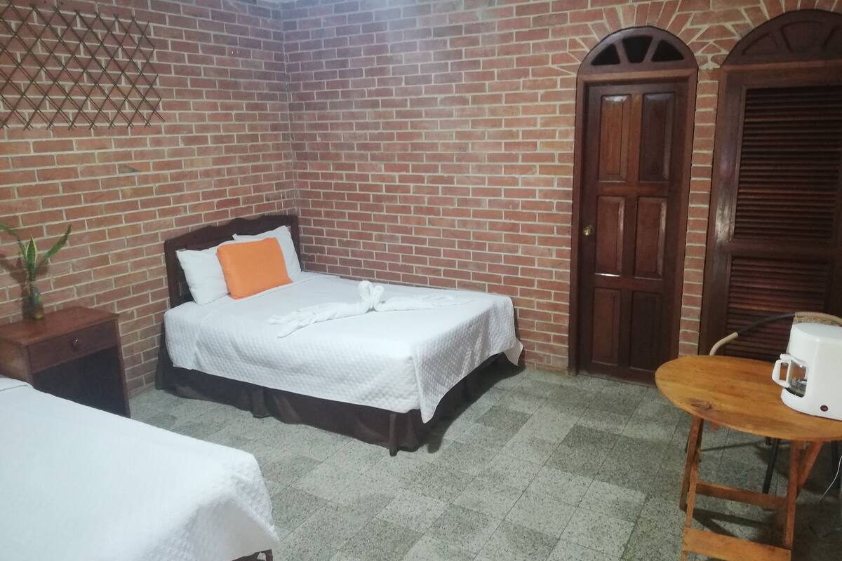 A hotel room with a brick wall, a bed with white bedding and an orange pillow, a wooden nightstand, a small table with a white coffee maker, and a door with a wooden frame.