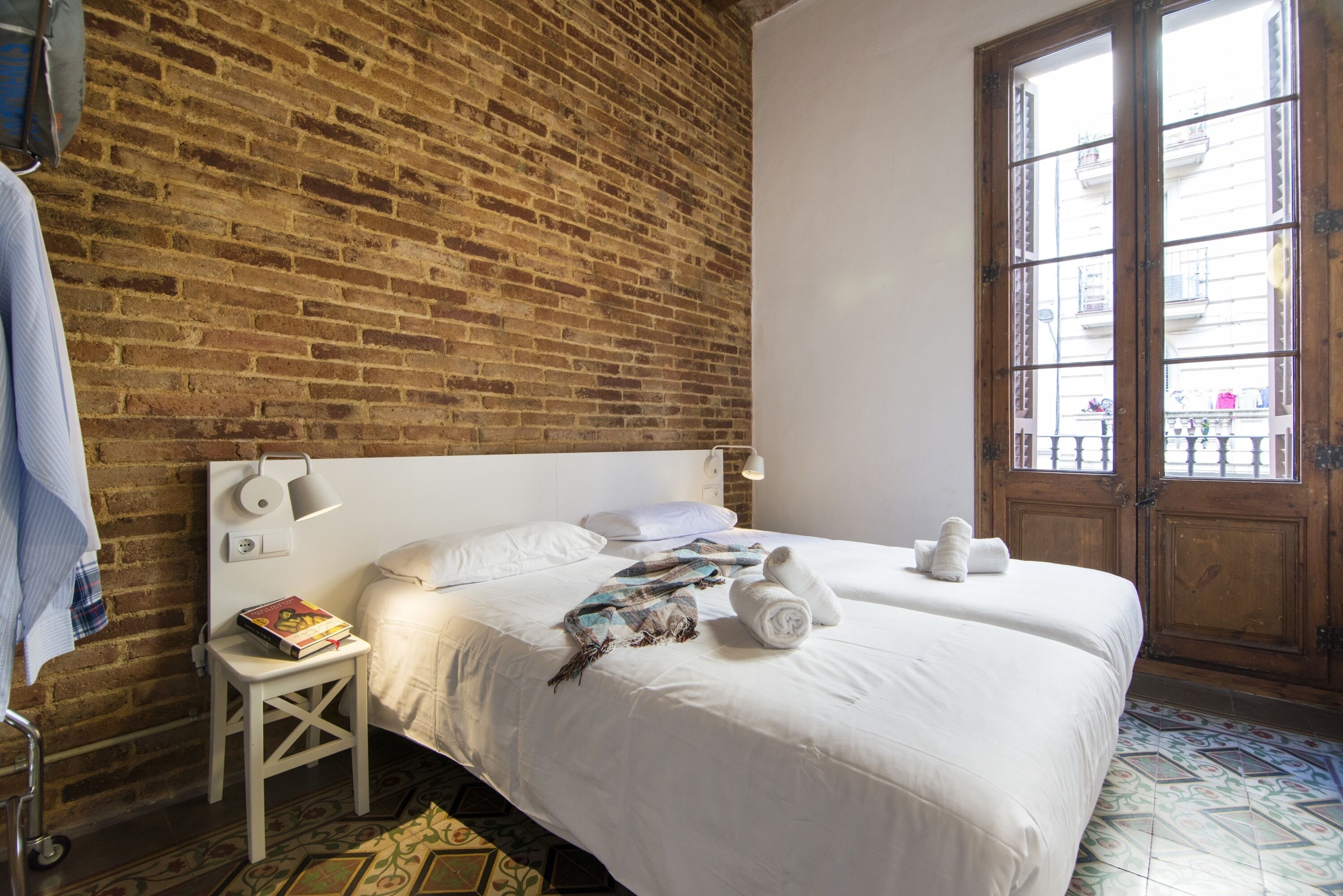 Habitación de hotel con pared de ladrillo, un ventanal con vistas a edificios, una cama con ropa blanca, una mesita con un libro y una toalla sobre la cama.