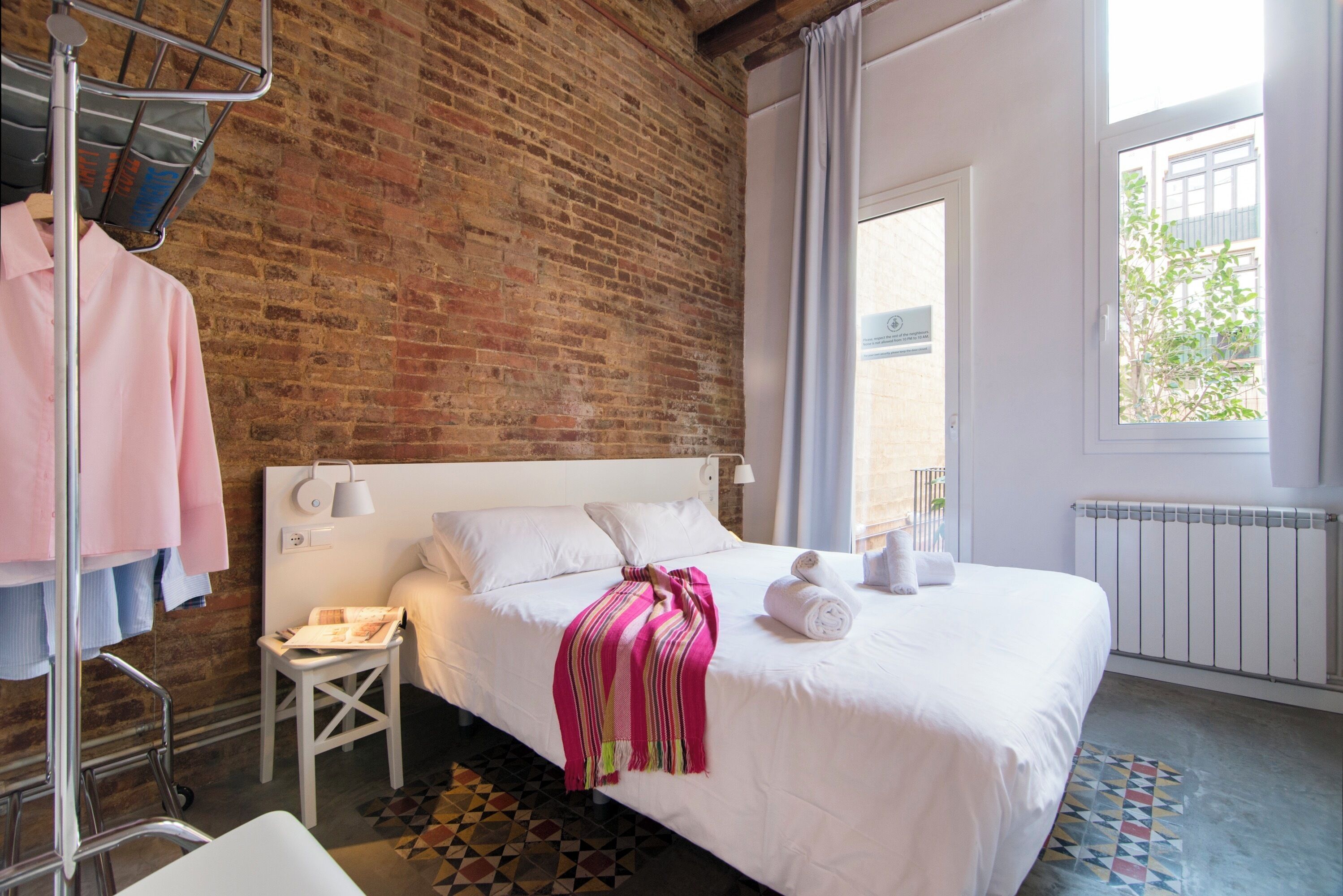 A hotel room with a brick wall, a large bed with white linens, a small white side table, and a window with curtains.