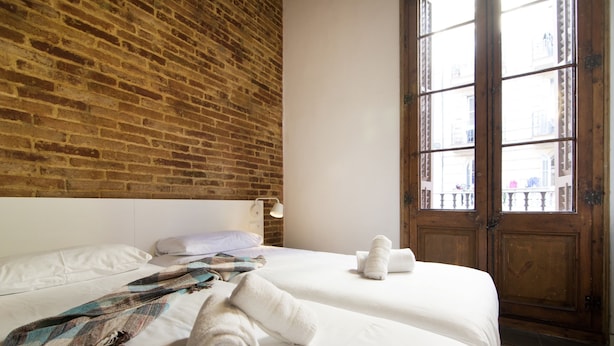 A hotel room with a brick wall, a large wooden door, and a bed with white linens and a plaid blanket. - Happy People Plaza España (Barcelona)