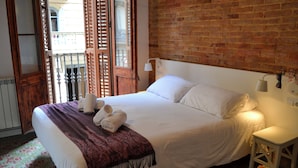 A neatly made bed with white linens and a purple throw blanket, a white bedside table with a lamp, and a brick wall in the background. - Happy People Plaza España (Barcelona)