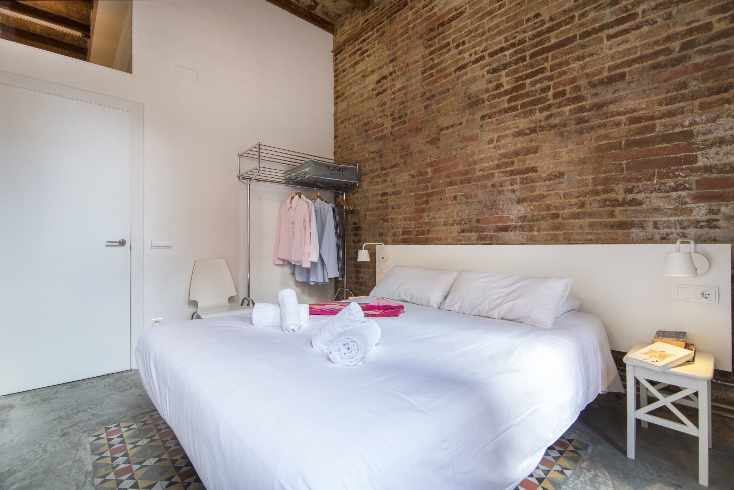 Une chambre d’hôtel avec un grand lit, un mur en briques, un porte-vêtements avec des serviettes et une table de chevet avec une lampe.