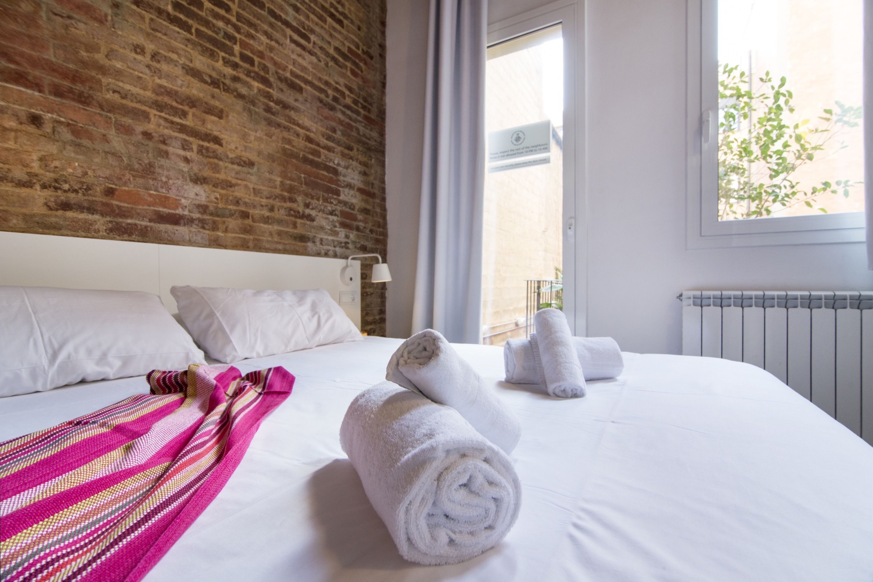 A hotel room with a brick wall, a large window, white bedding, and rolled towels on the bed.
