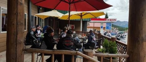 A wooden deck with outdoor seating, umbrellas, and a view of a gas station and mountains in the background.