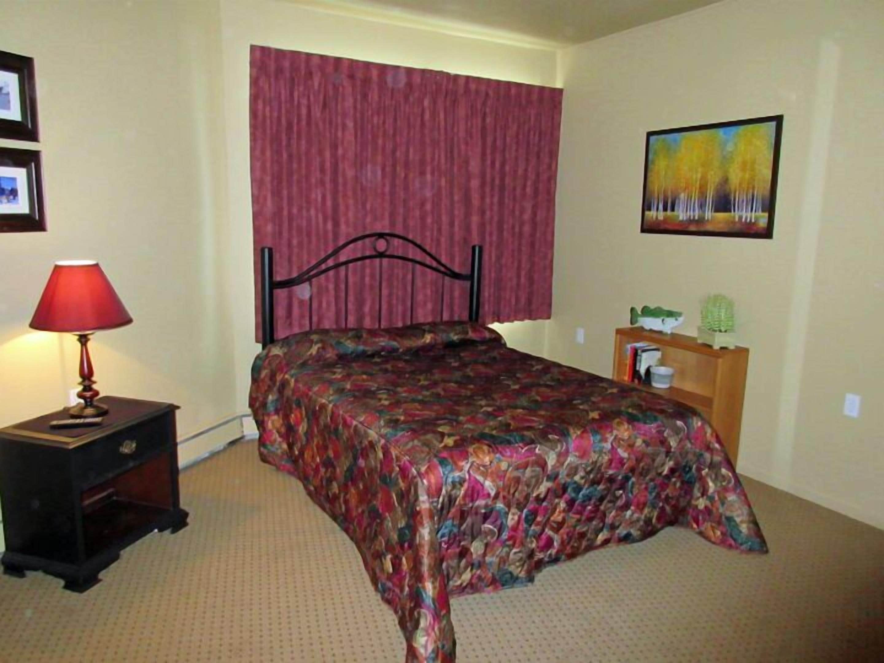 A bedroom with a bed, a nightstand with a lamp, a wooden dresser, and a painting on the wall.