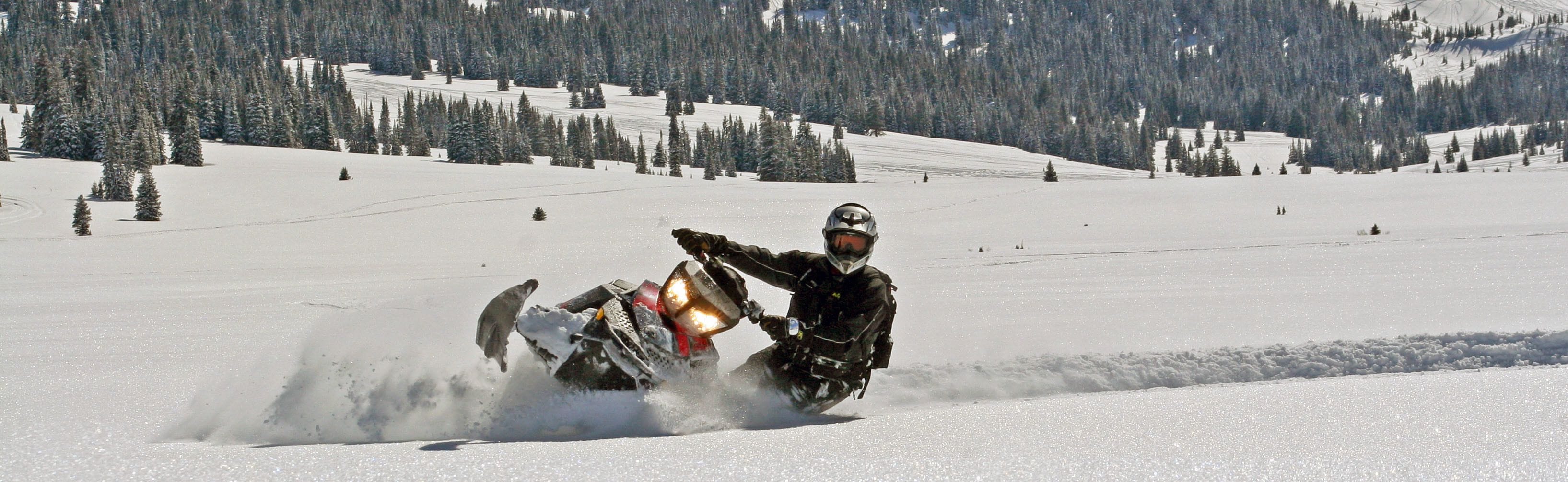 Um condutor de motos de neve em movimento numa paisagem coberta de neve, com árvores e céu limpo.