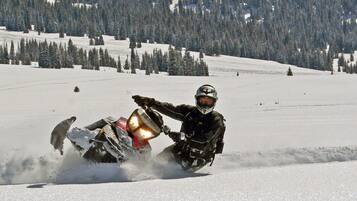 Um condutor de motos de neve em movimento numa paisagem coberta de neve, com árvores e céu limpo.