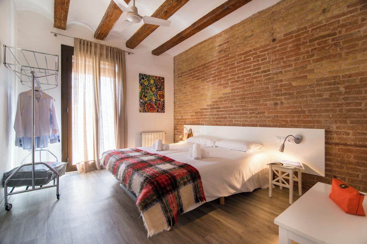 Een slaapkamer met een groot bed, een bakstenen muur, een plafondventilator, een raam met gordijnen en een radiator.