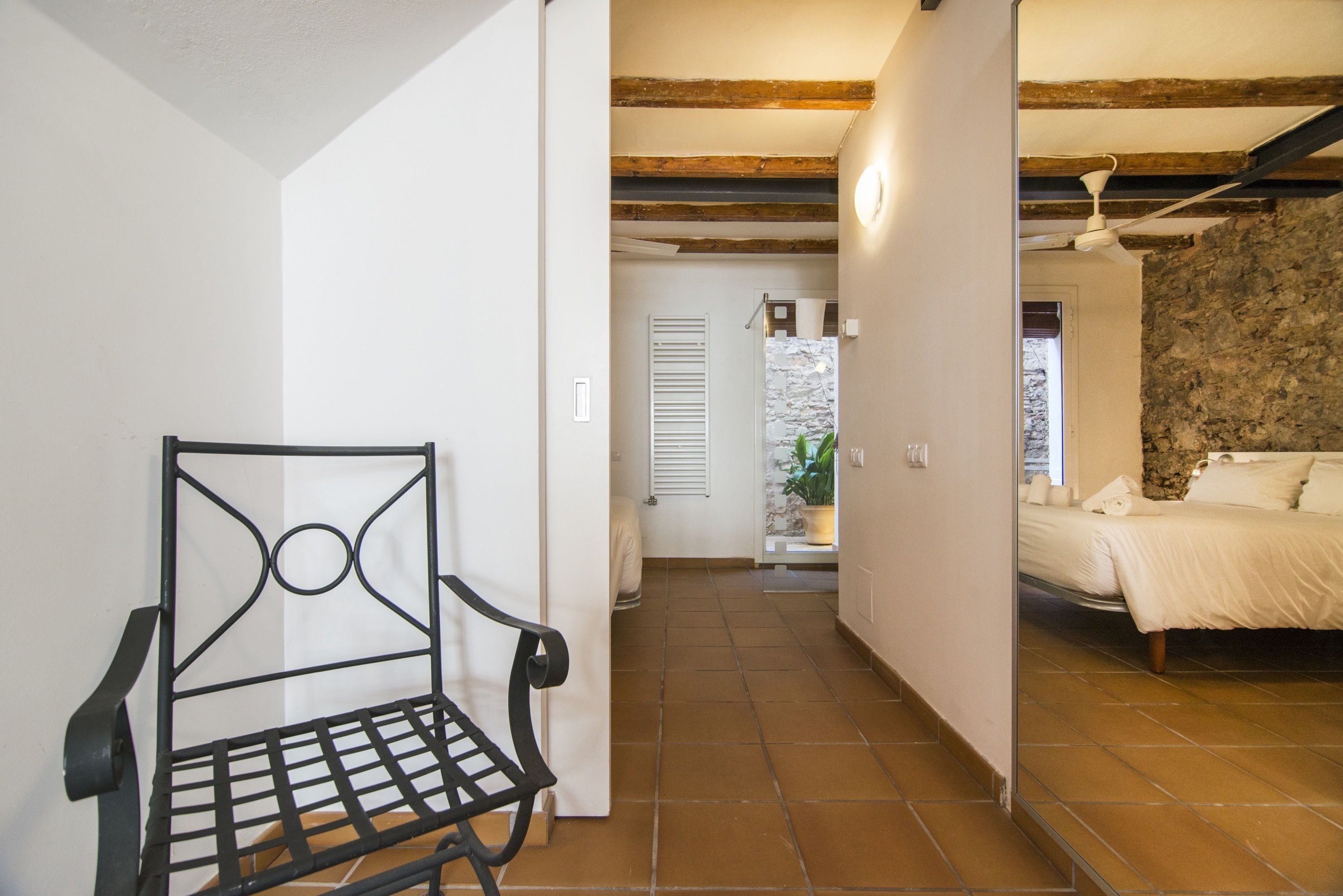Un couloir étroit avec une chaise en métal, un lit recouvert de draps blancs et un mur en pierre.