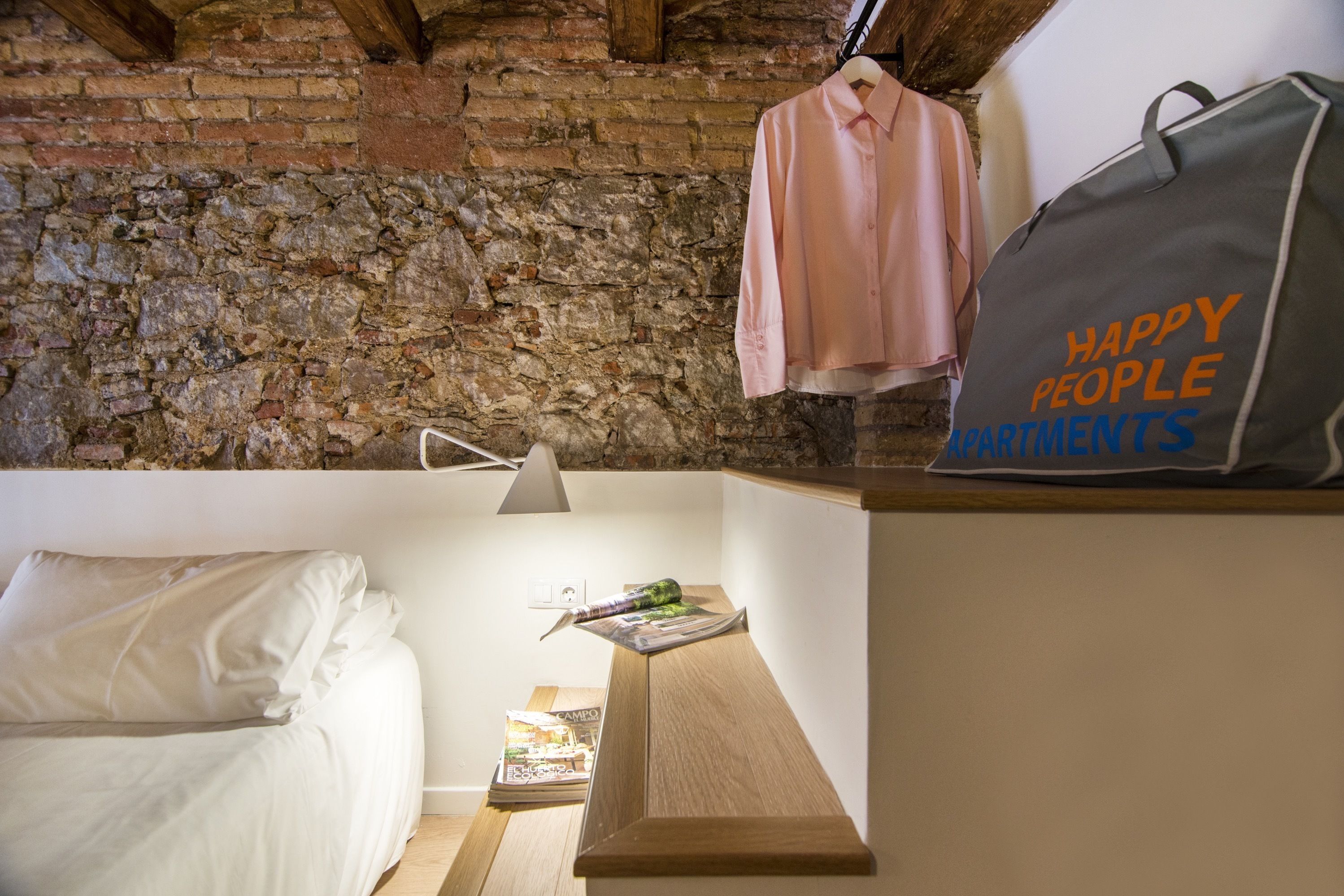 A bedroom with a brick wall, a bed, a hanging shirt, a magazine, and a bag with "HAPPY PEOPLE APARTMENTS" text.