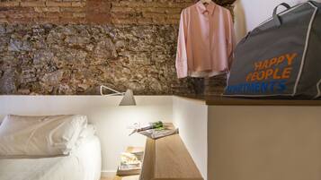 A bedroom with a brick wall, a bed, a hanging shirt, a magazine, and a bag with "HAPPY PEOPLE APARTMENTS" text.