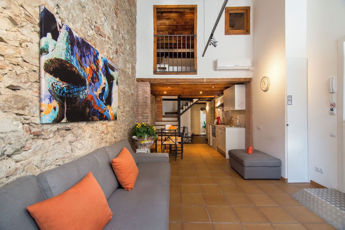 Een moderne woonkamer met een grijze bank, oranje kussens, een stenen muur en een kleurrijk abstract schilderij.