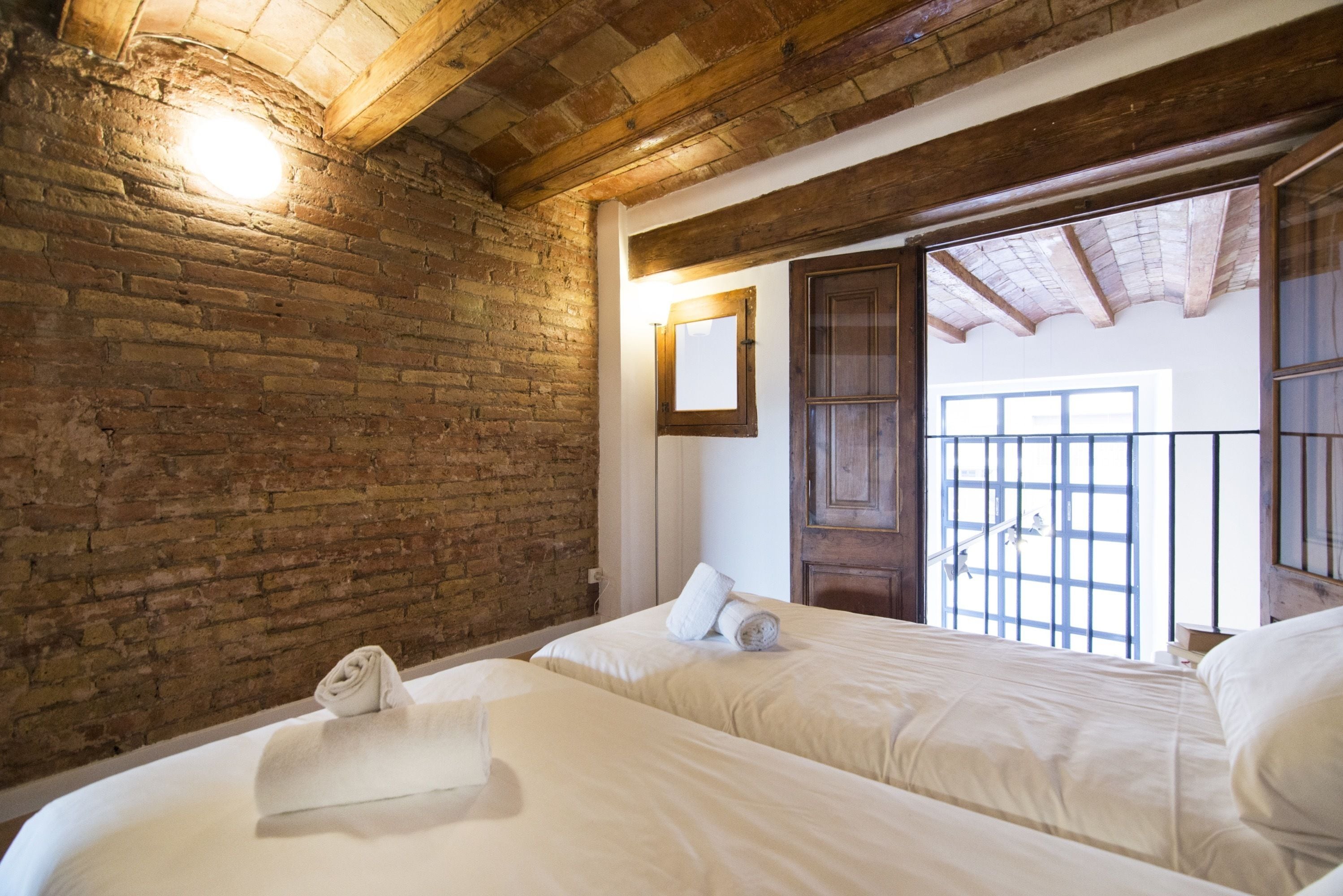 A room with two beds, a brick wall, wooden beams, a large window with bars, and a mirror.