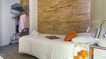 A neatly made bed with white bedding, a brick wall, a metal drying rack with clothes, and a small table with a glass jar and a book.