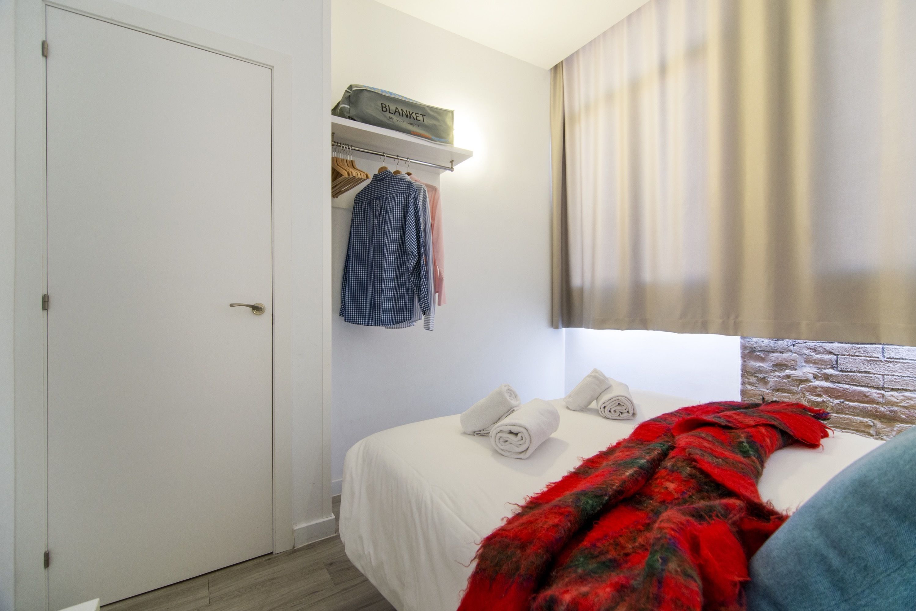 A bedroom with a bed, a wardrobe, a shelf with a blanket, and a window with curtains.