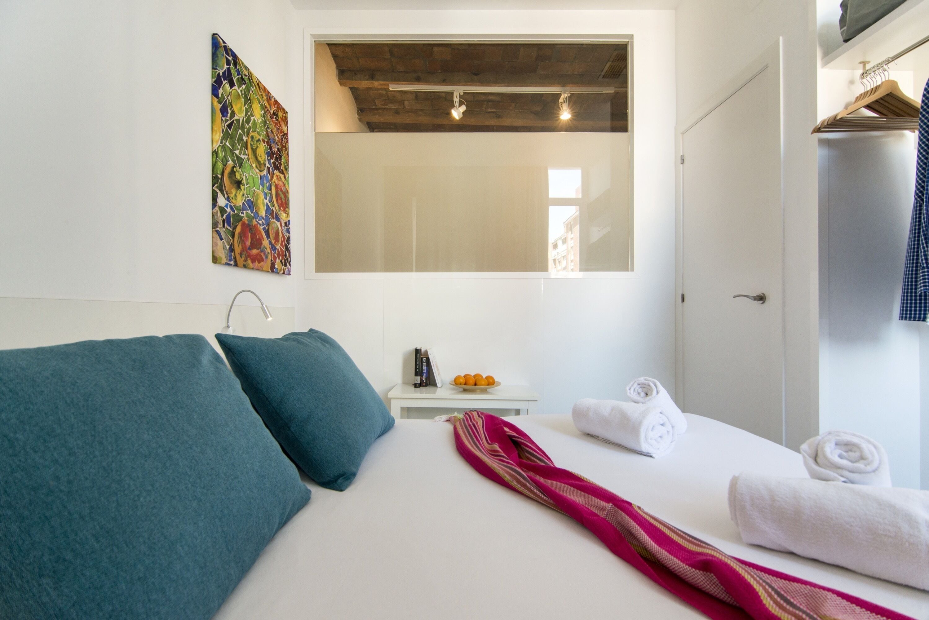Un dormitorio con cama, almohadas, una mesita de noche con frutero, una toalla y un perchero.