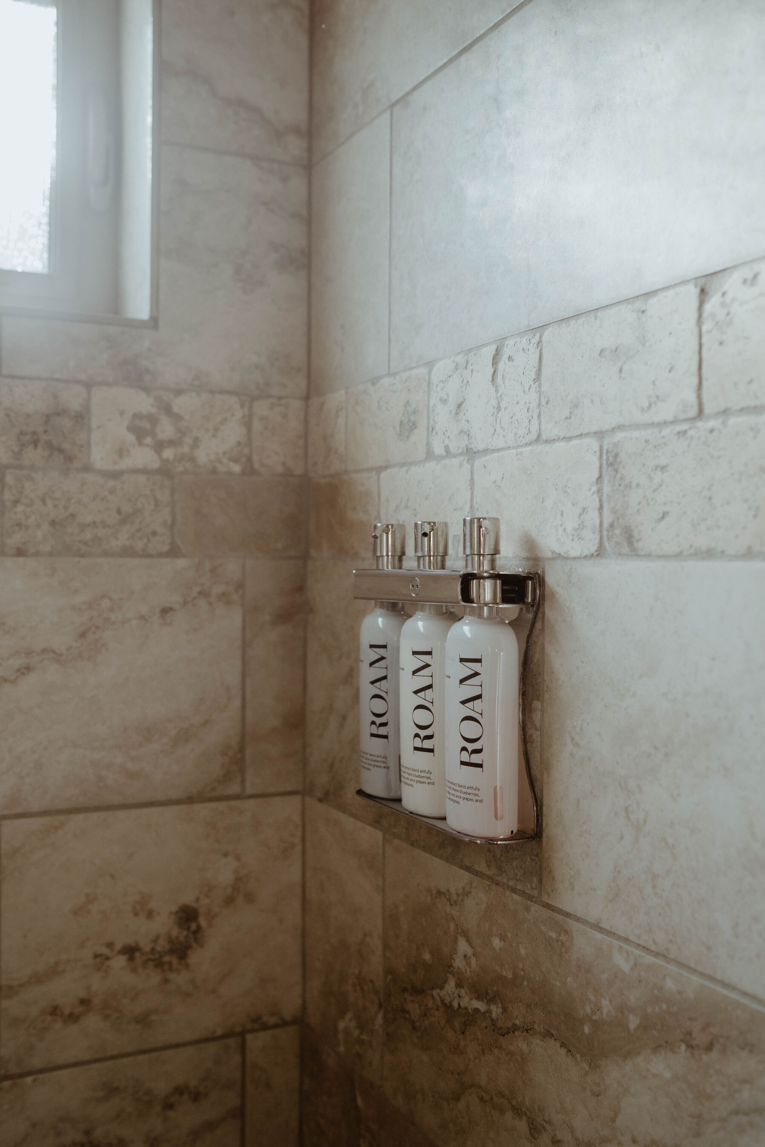 Une salle de bain avec un mur en marbre, une fenêtre et trois bouteilles étiquetées « CHAMBRE » sur une étagère.