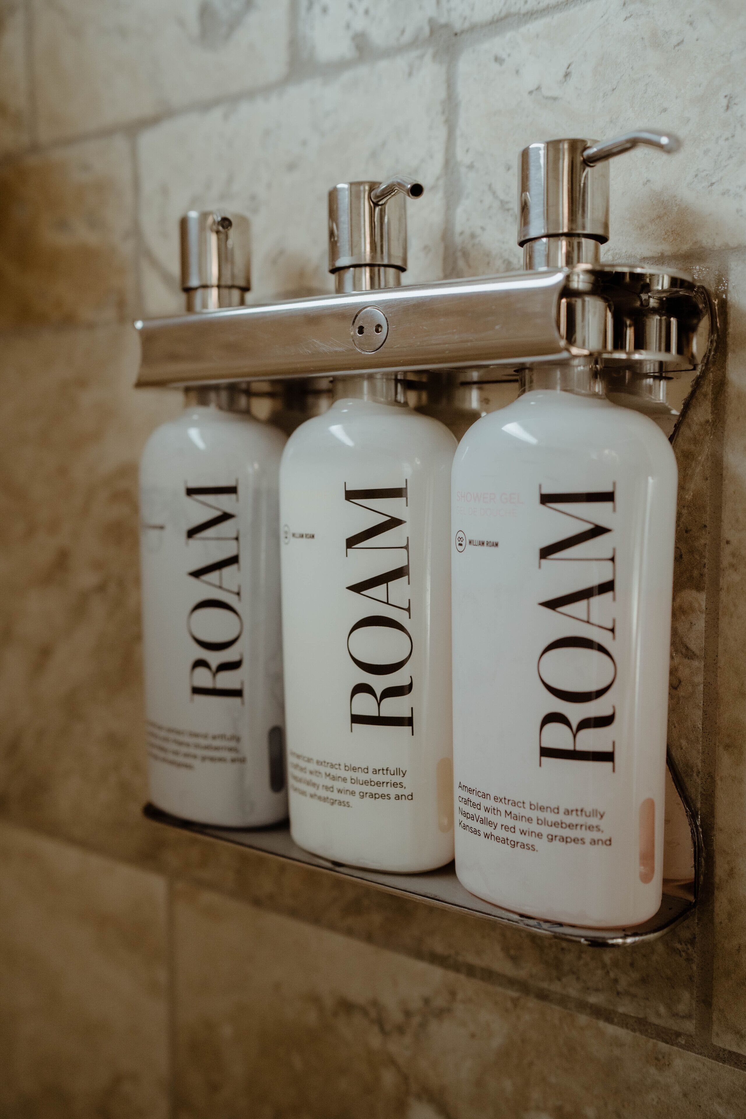 Three bottles of ROAM shower gel on a metal shelf against a tiled wall.