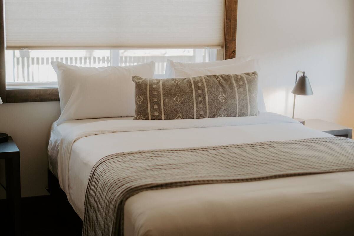 A neatly made bed with white and patterned pillows, a woven throw blanket, and a bedside lamp.