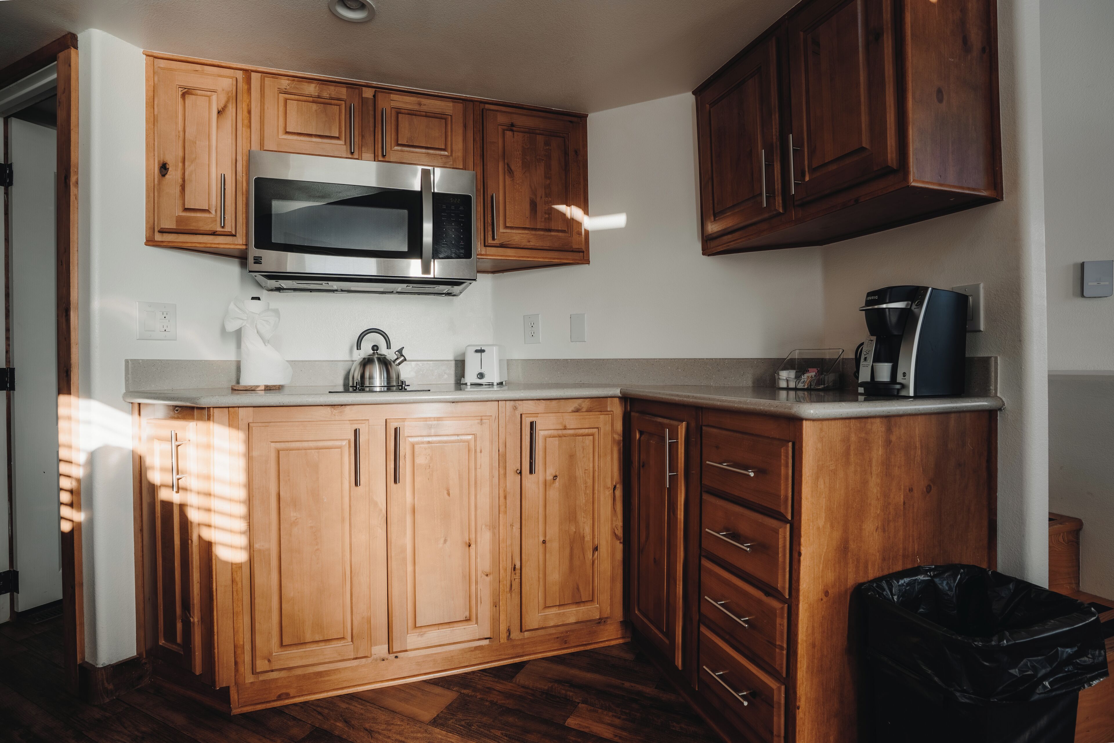 Una cocina con armarios de madera, un microondas de acero inoxidable, una cafetera y un cubo de basura.