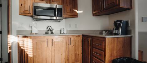 Una cocina con armarios de madera, un microondas de acero inoxidable, una cafetera y un cubo de basura.