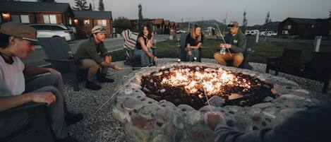 A group of people sitting around a stone fire pit in an outdoor setting with wooden cabins in the background.