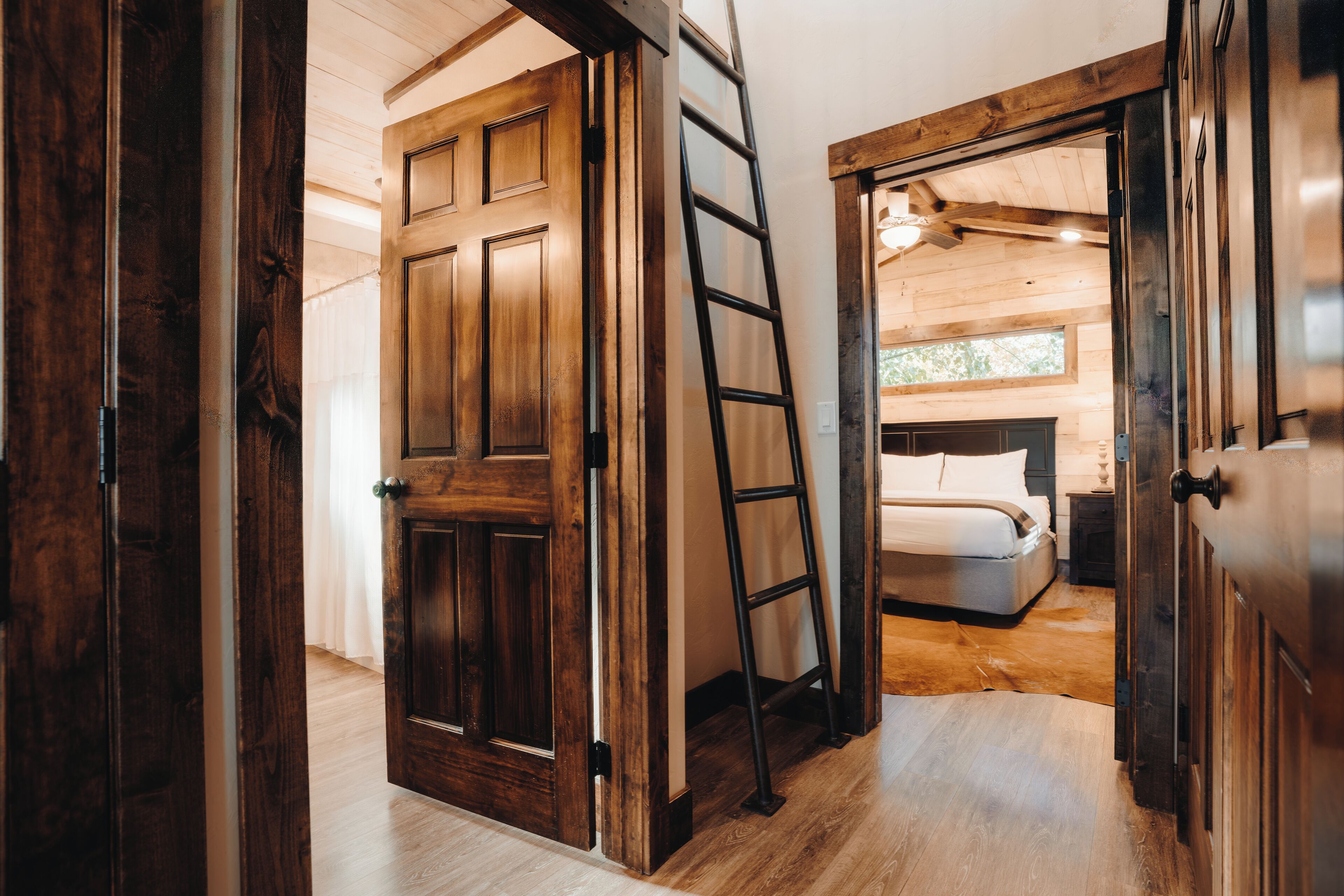 A wooden interior with a bed, a ladder, and a door leading to another room.