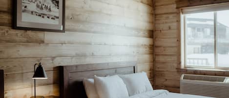 A wooden cabin room with a bed, a nightstand, a framed picture on the wall, and a window with a view of buildings.