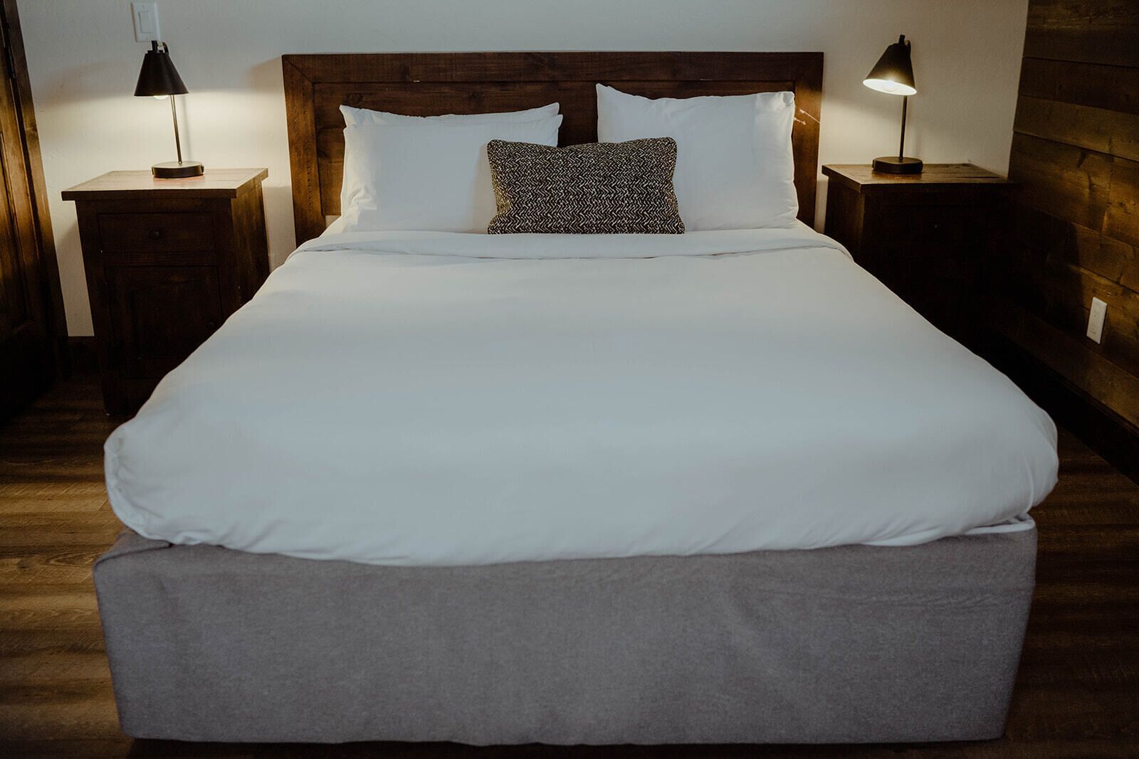 Ein ordentlich bezogenes Bett mit weißer Bettwäsche und grauem Fußteil, an dessen Seiten zwei Nachttische mit Lampen stehen, in einem Zimmer mit Holzboden und einem Holz-Kopfteil.