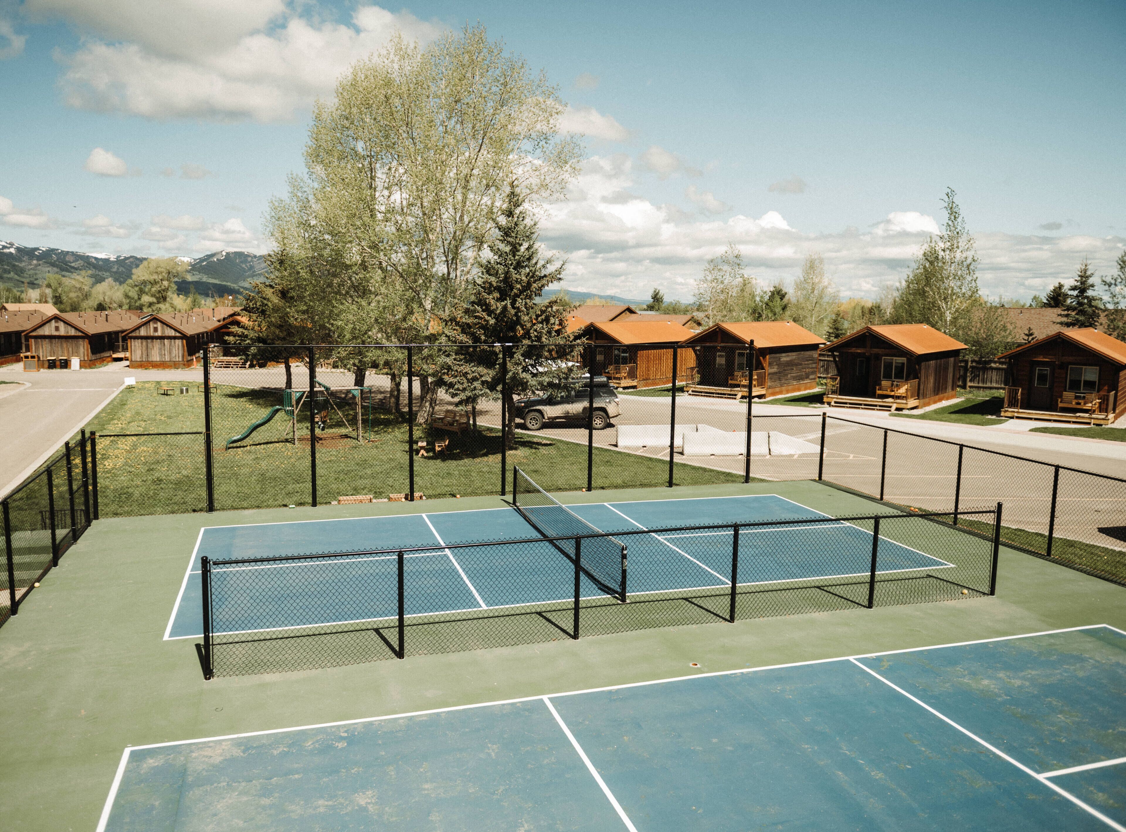 Un campo da tennis con rete, circondato da una recinzione, e casette di legno sullo sfondo.